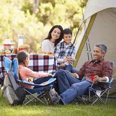 A family of four enjoys a picnic by their tent in a sunny, green park. They are smiling and relaxed, with food and drinks on a plaid tablecloth.